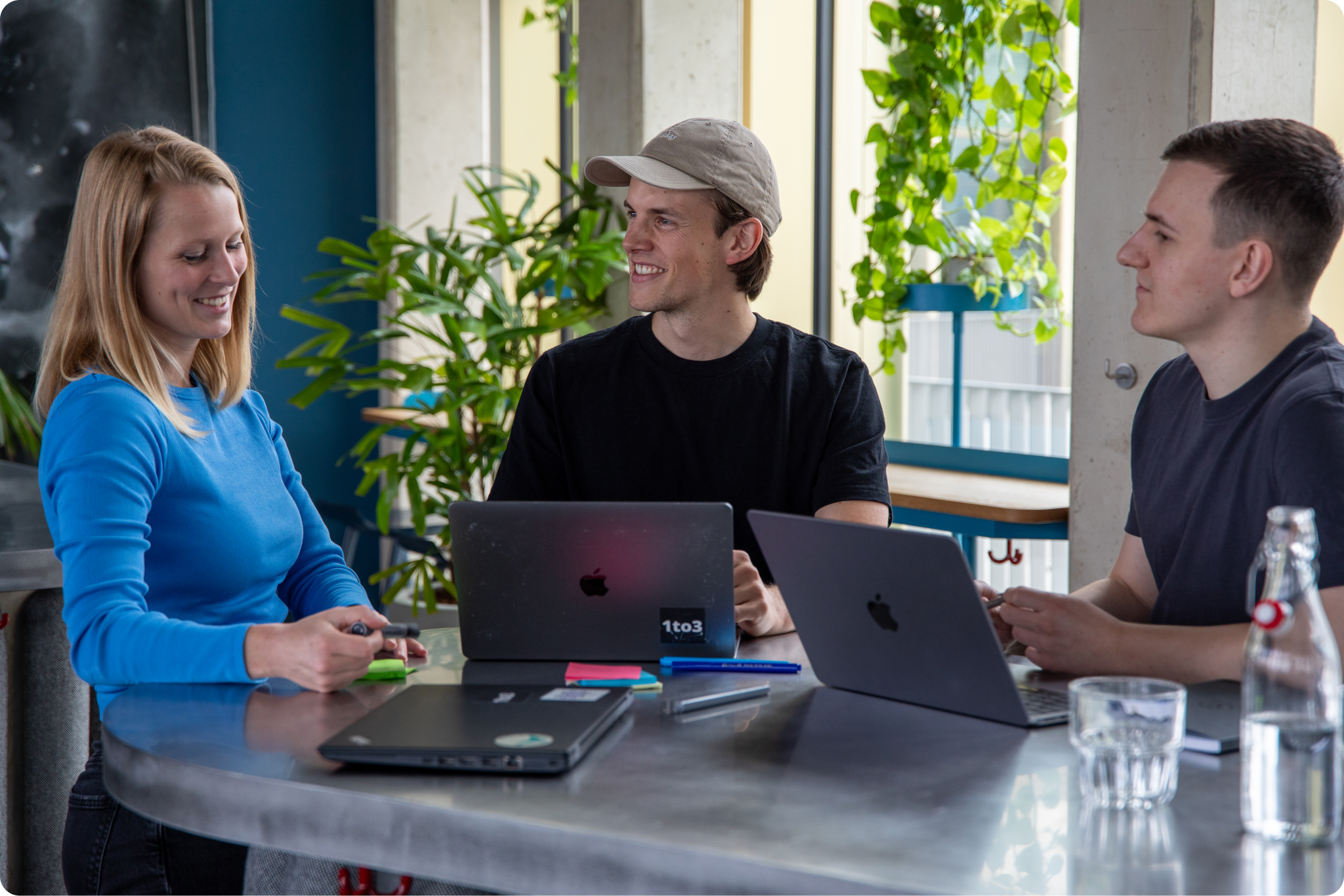 Drei junge Menschen sitzen mit Laptops an einem modernen Tisch, umgeben von Pflanzen, und führen ein entspanntes Gespräch in kreativer Atmosphäre.